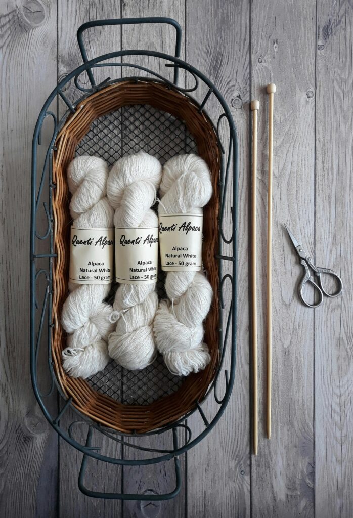 A flat lay of white alpaca yarn in a basket with knitting needles and scissors on wooden floor.